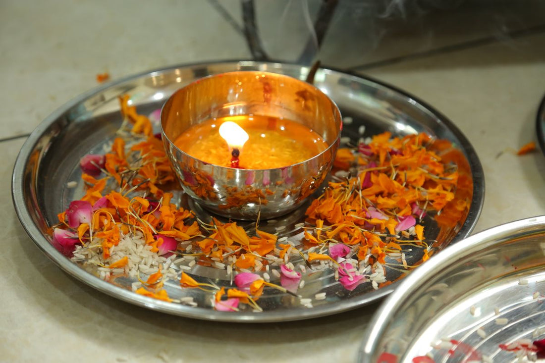 A lit diya on a metal puja plate surrounded by orange and pink flower petals